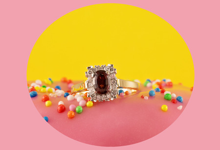 Garnet solitaire ring surrounded by diamonds set in a square shape with gold band on a pink iced cake with sprinkles, captured in a circle with a pink backgroundの写真素材