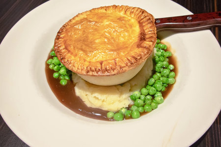 A steak pie with green peas and mashed potatoes and gravy served on a restaurant dinner plate