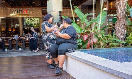 Brisbane, Queensland, Australia - 25th September 2019: Two overweight women drinking a milkshake each as they relax by the pool of a shopping centreのeditorial素材