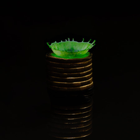 A green drop of milk splashed onto a stack of one dollar coins creates a crown shape with a water...