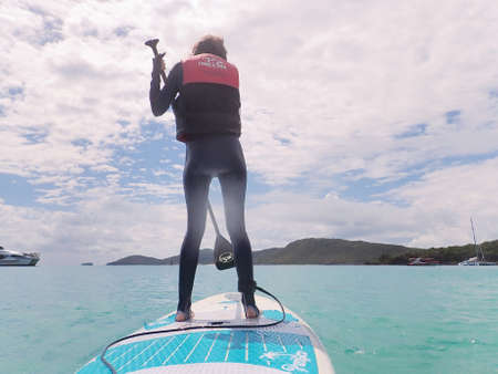 Whitehaven Beach, Whitsundays, Queensland, Australia - April 2021: Slim young woman in stinger suit standing on a surfboard paddle board with sun flareのeditorial素材