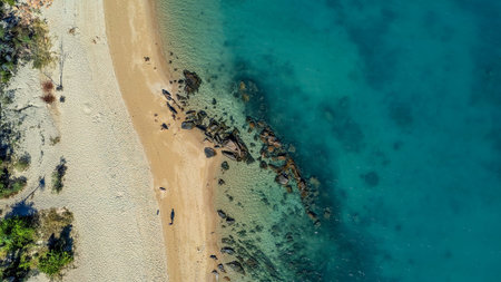 A man and his dog walking on the beach far below a drone flying overhead on a tropical beach at low tide with rocks showing out of the water and details on the seabed of aqua blue oceanの写真素材