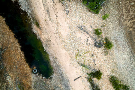 Looking down by drone into a pool of dark wire with a rubber car tire floating on the edge and a stoney shorelineの写真素材