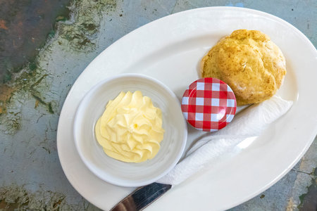 A scone with cream and a pot of jam on a white plate with cutlery