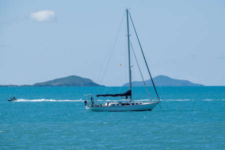 Airlie Beach, Queensland, Australia - December 2021: A jet ski powering back a luxury yacht with its sail down anchored offshore with island and mountain background against a blue skyのeditorial素材