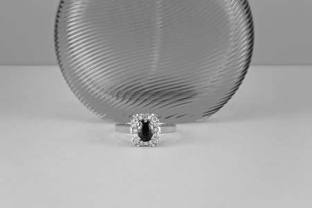 Garnet solitaire ring surrounded by diamonds in a square shape with gold band in front of a transparent perfume bottle isolated on a light surface.の写真素材