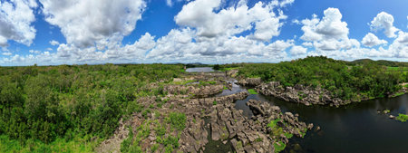 A panormama of a weir wall on a rock strewn river through bushland under a cloudy blue sky....