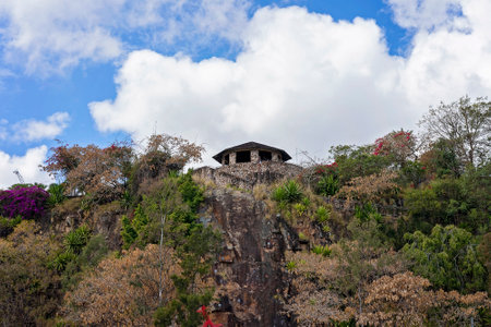 Old stone building on a cliff covered in vegetation and against a cloudy blue sky.の写真素材
