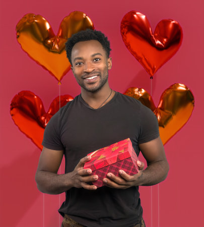 Radiant Valentine's Day moment: Handsome man holding a gift box surrounded by heart-shaped balloons on a cheerful pink background. Love, joy, and surprises!の写真素材
