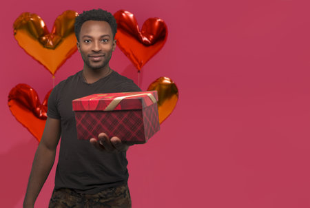 Radiant Valentine's Day moment with copy space: Handsome man holding a gift box surrounded by heart-shaped balloons on a cheerful pink background. Love, joy, and surprises!の写真素材