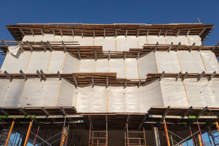 Facade of a modern building under construction featuring metal beams, scaffolding, and protective...