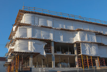 Contemporary building construction site wrapped with protective white sheets and scaffolding, clear blue sky background.の写真素材