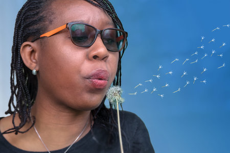 African woman with sunglasses blowing dandelion seeds, isolated on blue background. Perfect for...