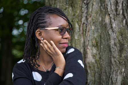 African woman wearing glasses and braided hair gently smiling outdoors, hand thoughtfully touching...
