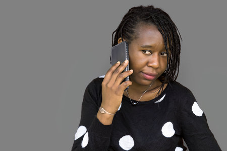 African woman with braided hair and polka dot sweater talking on a smartphone, isolated on gray background. Perfect for communication, technology, business, and lifestyle themes.の写真素材