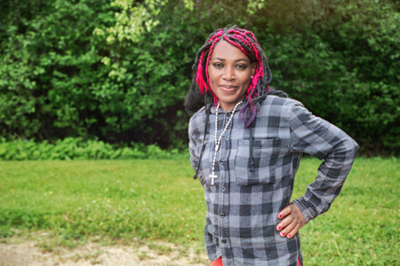 Confident young Black female with colorful protective hairstyle wearing plaid shirt outdoors,...
