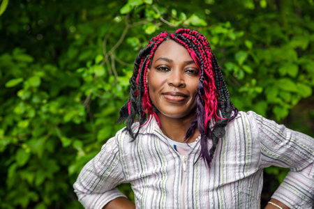 Smiling woman with colorful protective hairstyle in casual striped top, natural green background...