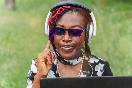 Confident young Black woman with glasses listening to music with headphones outdoors while using...