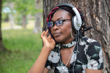 Black woman with glasses listening to audio content with headphones outdoors. Perfect for podcast,...