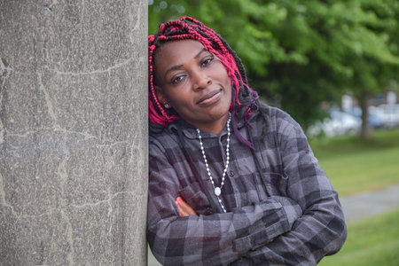 Confident mature African woman with arms crossed leaning against concrete column in park. Beautiful...