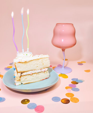 A Vanilla Birthday Cake with Lit Candles, A Pink Wine Glass and Confetti on a Pastel Pink Backgroundの写真素材