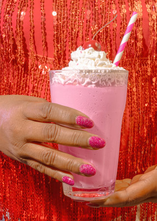 A Diverse African American Female Dressed in Red Sequins and Wearing Pink Nail Polish Holding a Pink Dessert Strawberry Milkshake with Whipped Creamの写真素材
