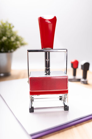 An automatic red, rubber date stamp placed on a paper folder. An office scenery. A composition of...