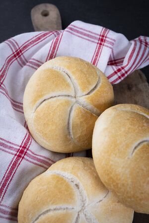 Rustic composition of traditionally baked, crusty Kaiser rolls. Bread rolls placed on a linen, checkered kitchen cloth and a wooden cutting board, on a dark background.の写真素材