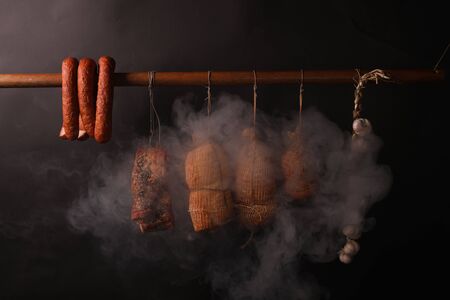 A composition of freshly smoked cold cuts hanging on strings. Traditional smokehouse with oak wood smoke. Freshly smoked pork sausages, hams and bacon in a smokehouse. Smoked pork meat.の写真素材