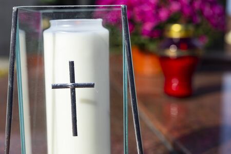 An elegant, glass, grave candle. All Saints' Day on a Christian cemetery. A close-up of a candle with a Christian cross. Granite tombstone decorated with chrysanthemums and candles in a background. の写真素材