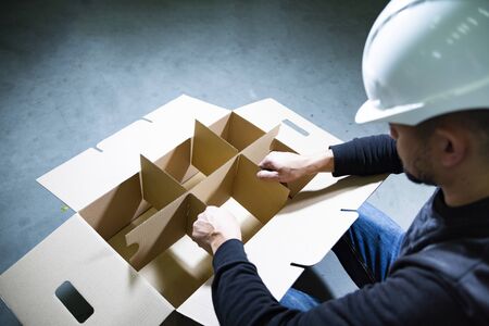 Top view of a man with white hard hat assembling a large, open cardboard box with compartments on the floor in a warehouse. Empty box on gray stone floor plates. A carton box for bottles with handles.の写真素材