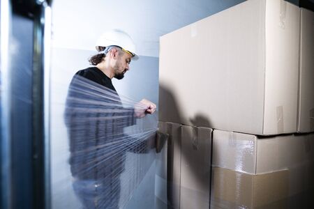 Wrapping pile of cardboard boxes in a stretch foil. A man packing cartons in a warehouse in a factory.の写真素材