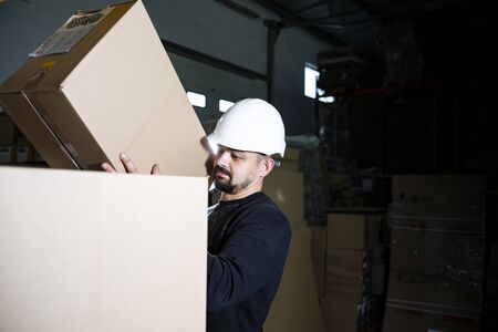 Storage work. Man with a hard hat placing carton box on a pile in a warehouse.の写真素材