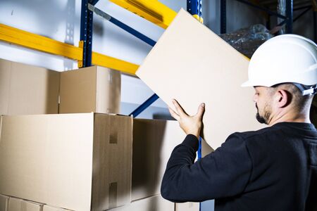 Warehouse worker. Man with a hard hat placing a carton box on a pile on a storage shelf.の写真素材