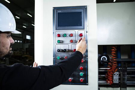 Man in a hard hat operates printers in a production hall. Inputing printing settings. Cardboard boxes factoryの写真素材