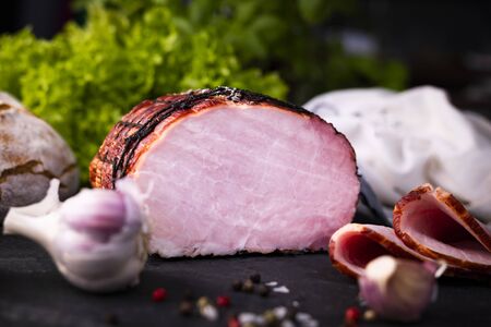 A piece of smoked, homemade, boneless ham in netting in composition with garlic, peppercorns and crunchy bread, on a stony, kitchen countertop. Traditional meat products.の写真素材