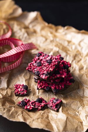 Chocolate pieces with freeze-dried strawberries and blueberries on a paper with a decorative red checkered ribbon. Moody, appetizing composition.の写真素材