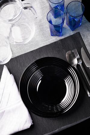 Modern tableware for one person. A top view of black plates, cutlery, an elegant linen napkin, glasses and jug on a gray tablecloth and a black, woven, rectangular placemat.の写真素材