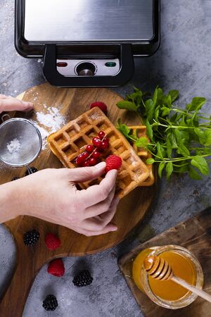 Top view on a male hands decorating freshly baked, tasty, golden waffles with powdered sugar, fresh berries and honey on wooden cutting board, next to professional waffle iron. Belgium waffles.の写真素材