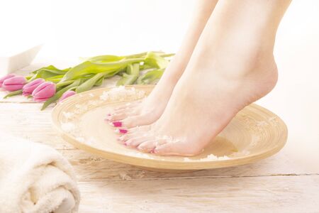 Perfect pedicure. A young woman taking warm SPA footbath with special salts. Body care, foot relaxing in cozy place with wood floor decorated with tulips. Wellness, day SPA, beauty salon. Women's Dayの写真素材
