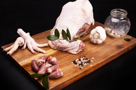 Poultry meat and giblets composition. Raw chicken carcass, hearts and feet on a wooden cutting board on a black background. Chicken broth portion decorated with bay leaves, garlic, salt and pepper.の写真素材