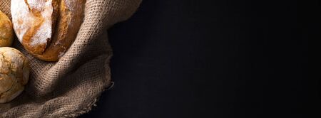 Breadstuff on jute fabric and a black background. Loaf of freshly baked, crispy, handmade, country bread and kaiser bread rolls. Baked goods, wide banner with a copyspace, text space.の写真素材