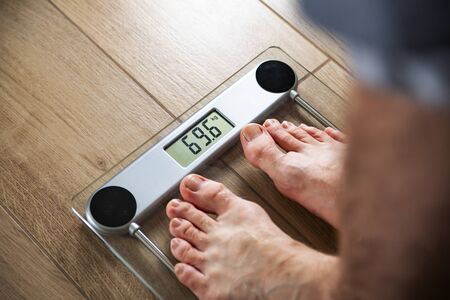 Close-up of a male feet of an young fit man standing on electronic scale on a wooden-like, tile floor in a bathroom. Weight control. An underweight, slimming, weight gain, loss. Body balance.の写真素材