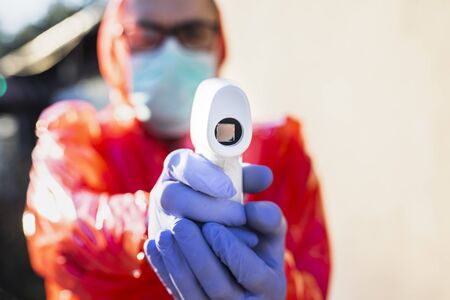Man in protective suit, gloves, glasses, surgical mask, controlling body temperature of drivers with an infrared forehead thermometer, outdoor on a sunny day.の写真素材