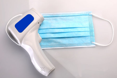 A one-use sterile surgical mask and an infrared forehead thermometer, isolated on a white...