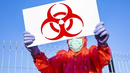 A man in a protective suit, wearing nitrile blue single use gloves and a surgical mask, standing outdoor with biohazard information board on a sunny day.の写真素材