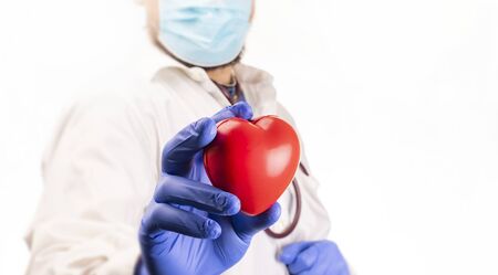 A doctor with a surgical mask and blue, protective nitrile gloves, holding a red heart in his hand, on a white background. A healthcare concept. A medical check-up.の写真素材