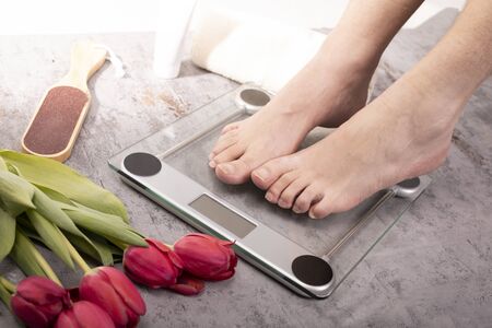 Healthy feet of a young fit man standing on electronic scale on a gray stony floor in a cozy bathroom, decorated with red tulips. Weight control. Body harmony, balance, relaxの写真素材