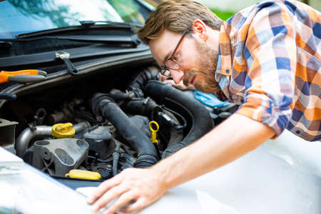 Young man with beard and glasses, looking for breakdown causes under the hood of the car. Fixing a car on a sunny day. Self auto repair. A car mechanics. Fixing a vehicle failure. Automobile service.の写真素材