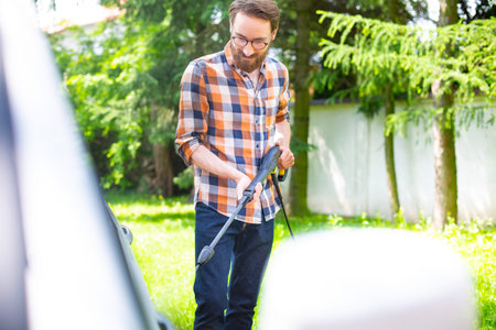 Handsome bearded man cleaning a car in the garden with a high pressure washer.